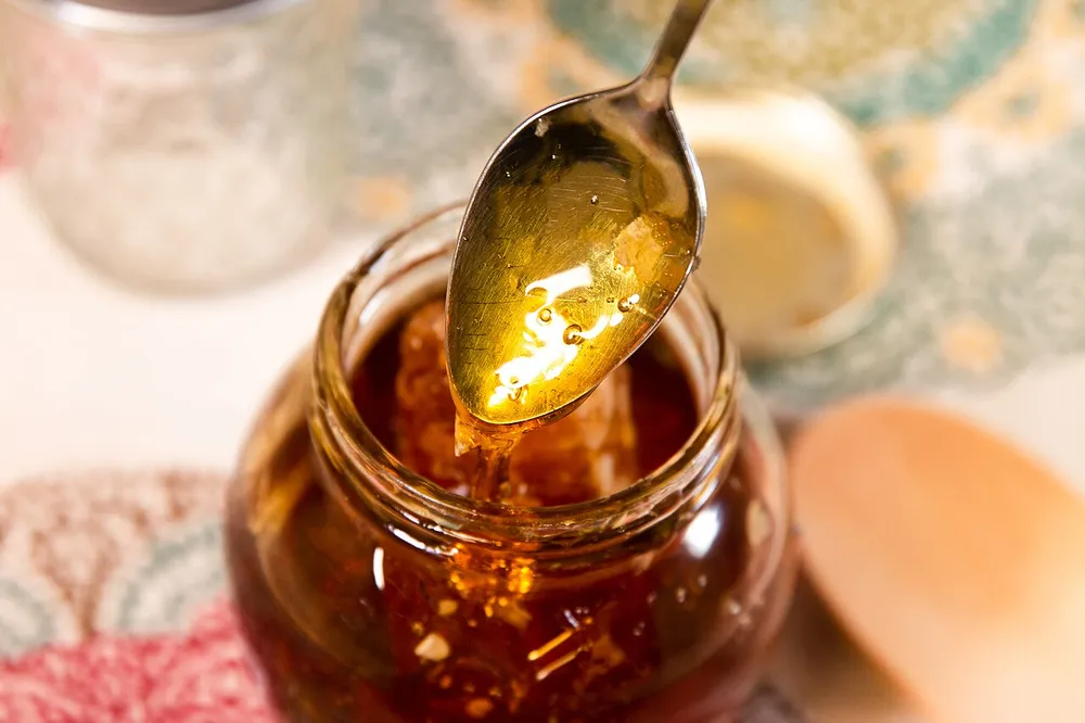 Ein kleines Glas Honig neben einem Stück Bienenwabe auf hellem Untergrund