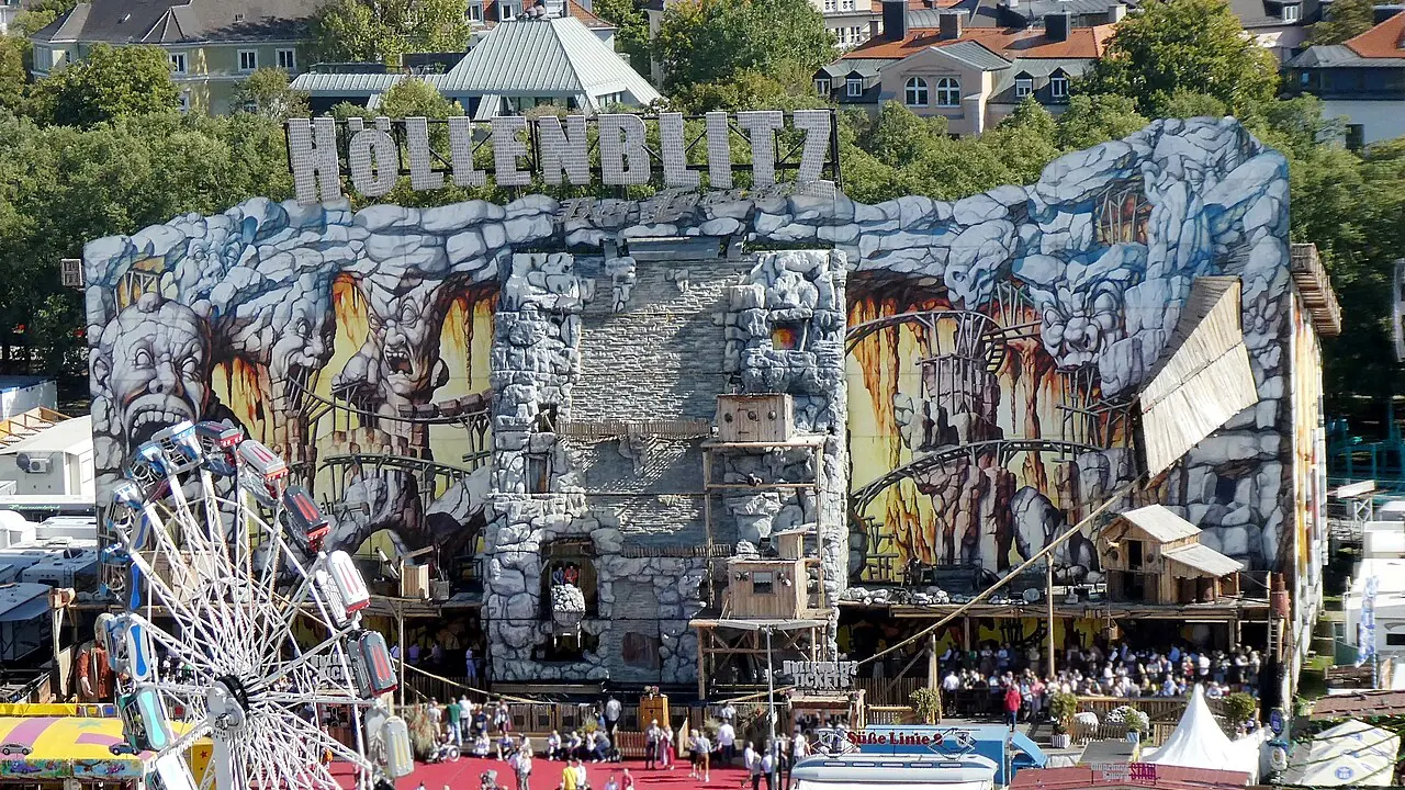 Blick über das Oktoberfestgelände auf der Münchner Theresienwiese: die bemalte Fassade der Achterbahn Höllenblitz, davor Besucher auf der Budengasse und ein Kettenkarussell