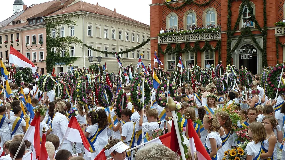 Kinder in weißer Kleidung mit blau-gelben Schärpen tragen Blumenkränze und Fahnen beim Festzug des Kamenzer Forstfestes über den Markt