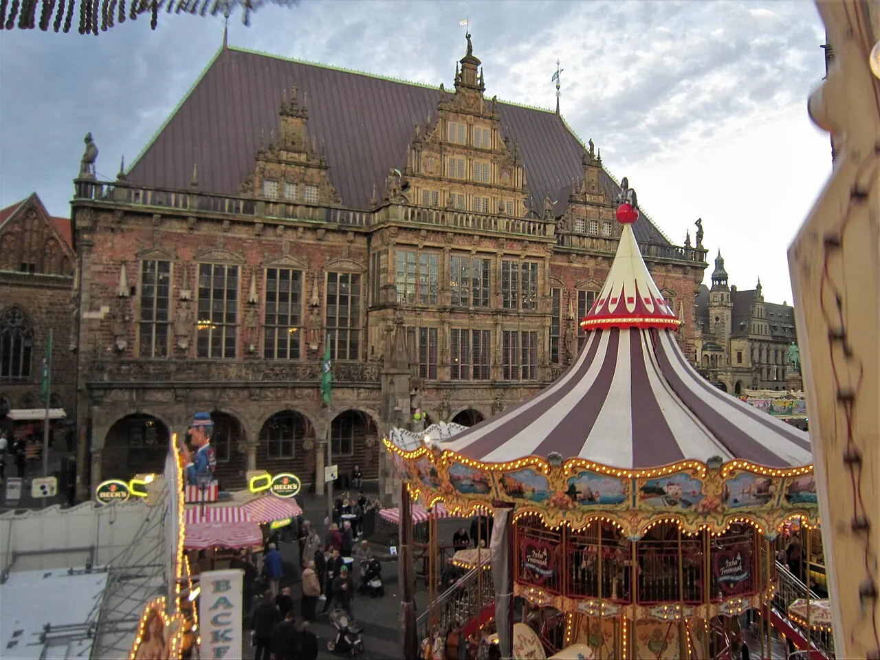 Blick aus dem Riesenrad auf den Kleinen Freimarkt auf dem Bremer Marktplatz: vorn ein beleuchtetes nostalgisches Karussell mit rot-weiß gestreiftem Dach, dahinter das Bremer Rathaus mit seiner Renaissancefassade