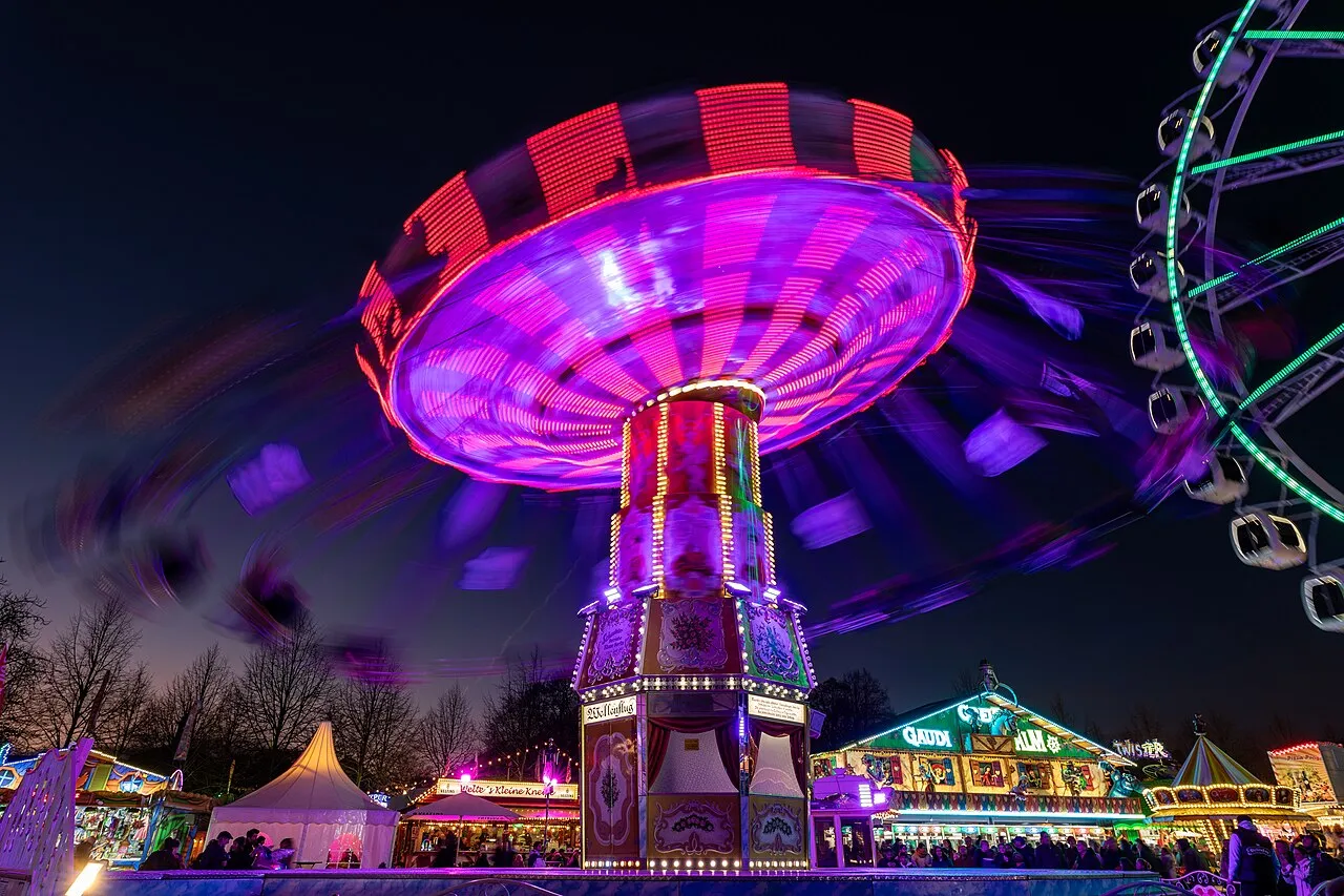 Der beleuchtete Wellenflug beim Herbstsend auf dem Münsteraner Schlossplatz am Abend: die Gondeln drehen sich in Bewegungsunschärfe unter einem pink und rot leuchtenden Dach, rechts ein grün beleuchtetes Riesenrad, unten Buden und Besucher